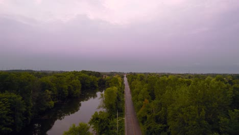 Toma-Aérea-De-Seguimiento-De-Drones-De-La-Carretera-Rural-Al-Atardecer-Junto-Al-Estanque-Y-La-Zona-Boscosa-Con-Un-Paisaje-Escénico-De-Cielos-Nublados