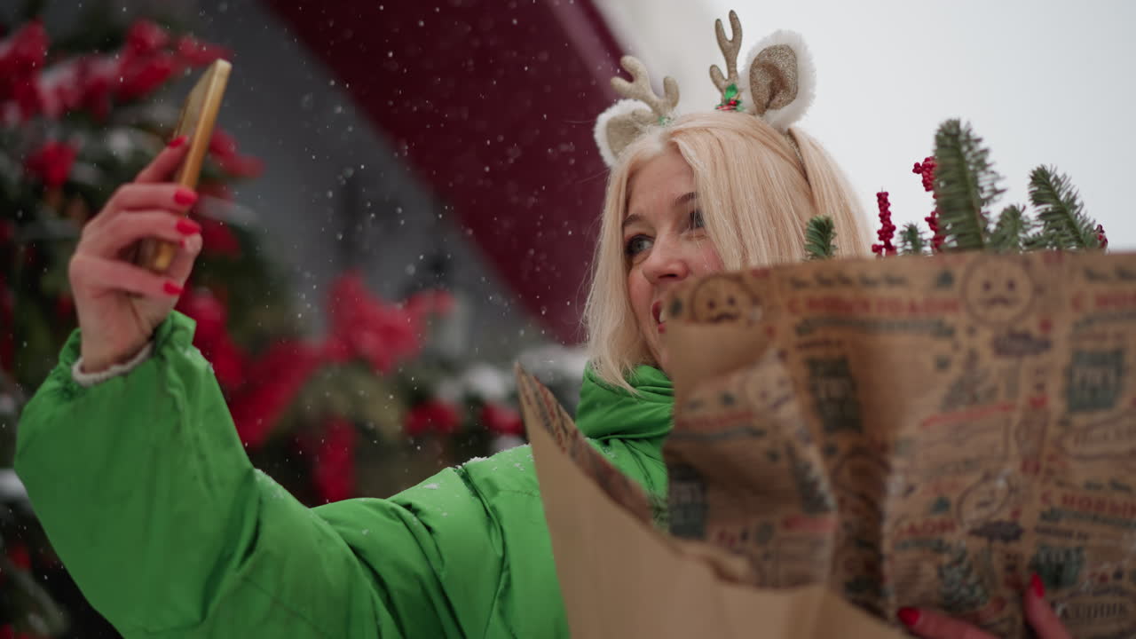 Mulher branca segurando presente de Natal em transmissão ao vivo em rua nevada, vestindo casaco acolchoado verde e tiara de rena, mostrando buquê de papel impresso e sorrindo para a câmera do telefone como vlogger e doadora de presentes.