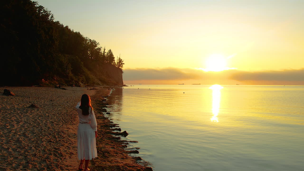 una chica con un vestido blanco está de pie junto al mar y mira fijamente el sol naranja naciente