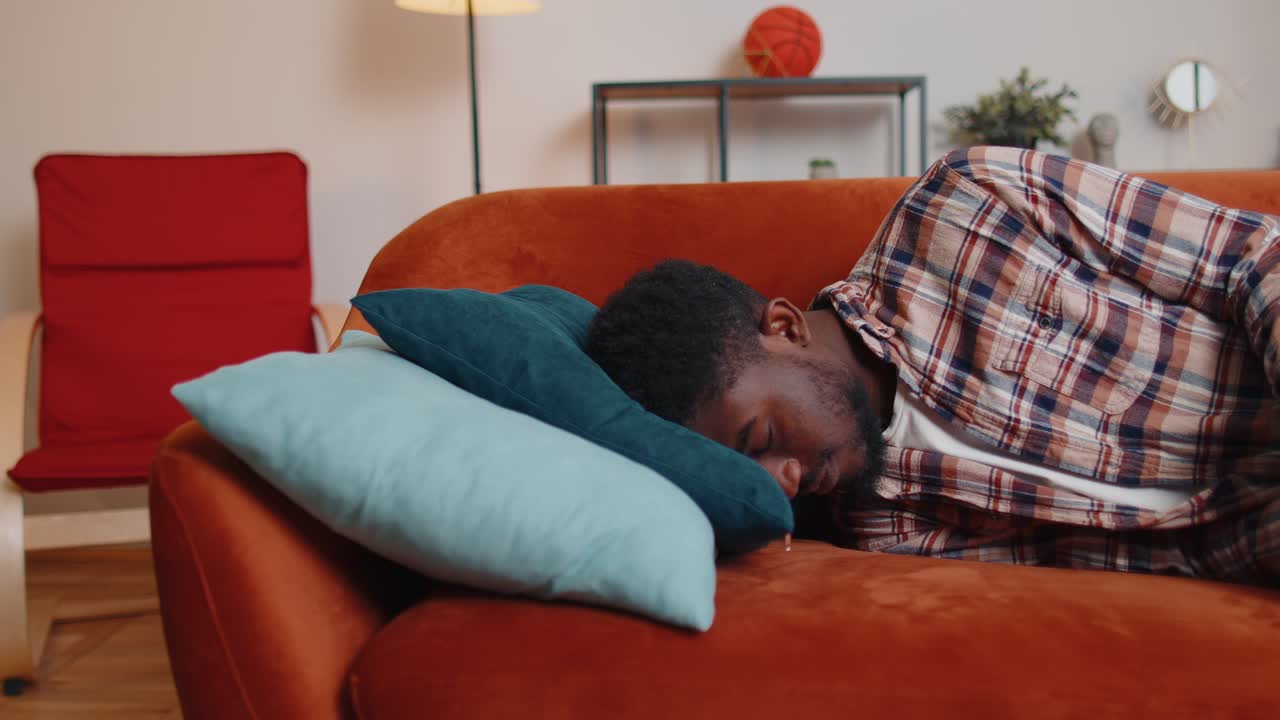 Homem afro-americano exausto e sobrecarregado adormecendo no sofá depois da festa, cansado, sobrecarregado.
