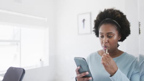 African-american-woman-brushing-teeth-and-using-smartphone-at-bathroom-mirror,-slow-motion