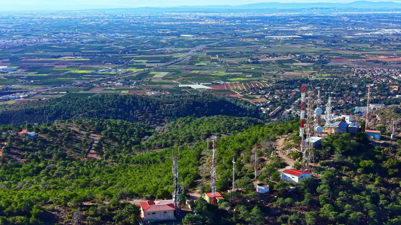 Vista di tralicci per telecomunicazioni su una montagna che si affaccia su un paesaggio di campi e strade in lontananza, in pieno giorno.