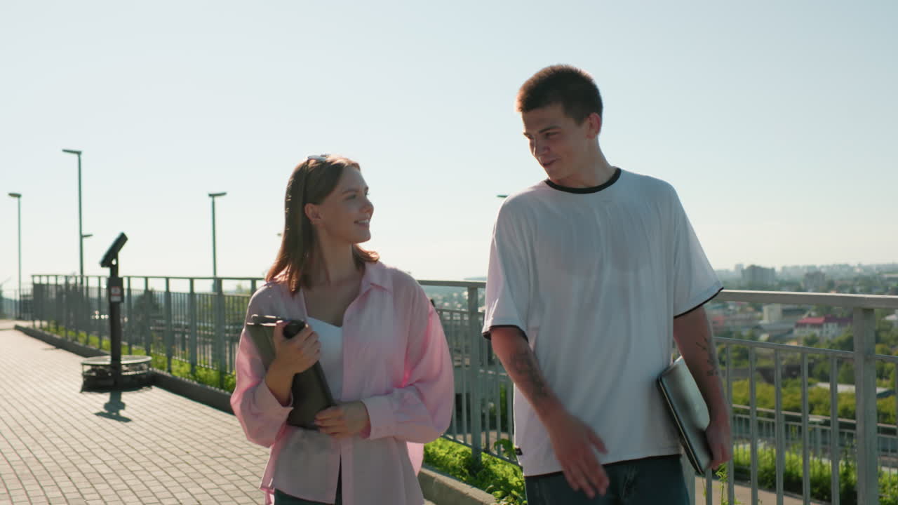 철의 레일링 근처에서 걸어가는 친구들, 문신한 팔을 가진 남자가 노트북을 들고 웃는 여자와 따뜻하게 대화하고, 은 하늘 아래 녹색과 넓은 도시 풍경을 보여주는 배경