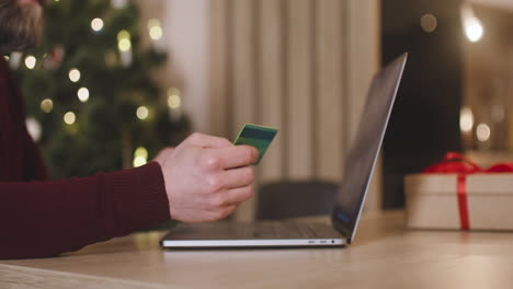 vista lateral de las manos de un hombre usando una laptop y sosteniendo una tarjeta de crédito sentada en una mesa cerca de un regalo en una habitación decorada con un árbol de navidad