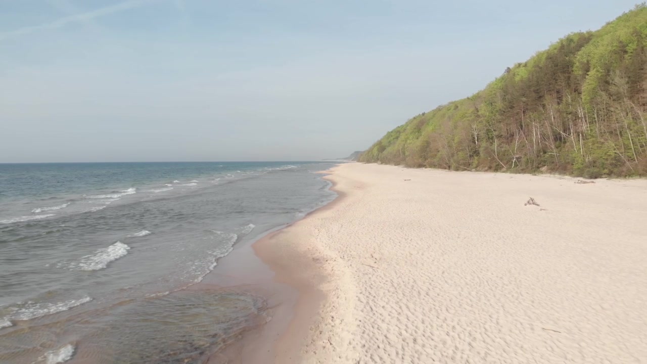 Ujęcie z drona płynnie cofające się nad spokojną piaszczystą plażą w Poddąbiu na polskim wybrzeżu Bałtyku, z delikatnymi falami, jasnymi wydmami i zalesionymi klifami w jasnym świetle dziennym.
