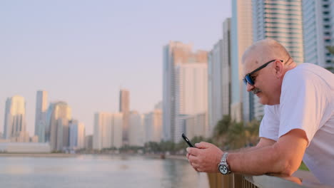 Happy-handsome-senior-man-in-sunglasses-and-white-t-shirt-writes-SMS-on-smartphone-standing-on-the-waterfront-in-summer-against-the-city-and-buildings