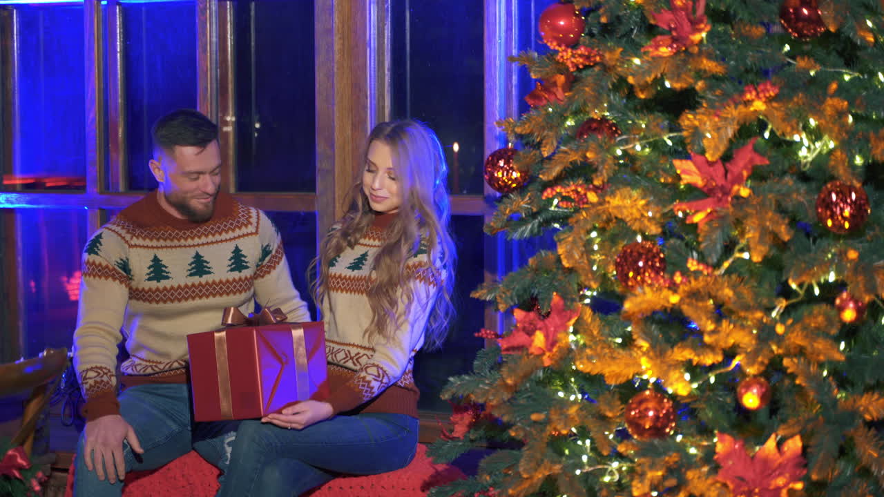 Happy romantic couple is celebrating Christmas or New Year together. Woman is receiving gift. Male and female sitting near Christmas tree.