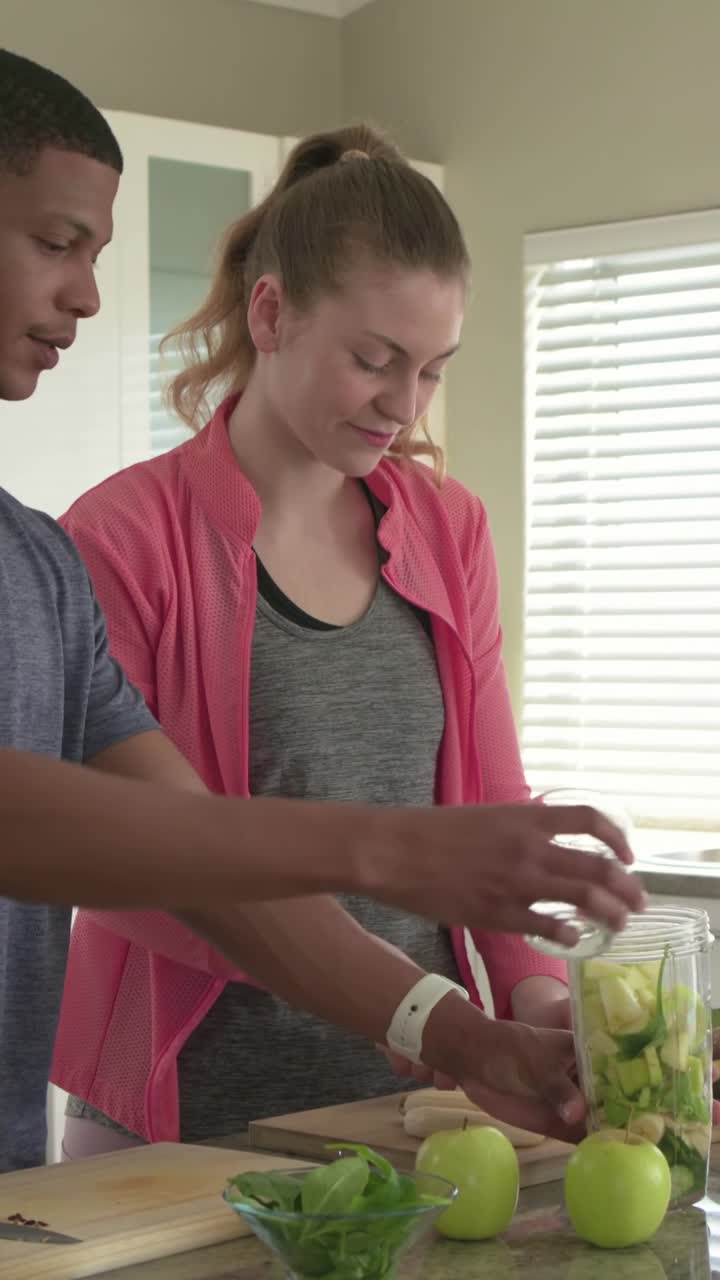 Video verticale: Donna in giacca rosa che prepara un frullato verde in cucina, con un uomo che si unisce e versa.