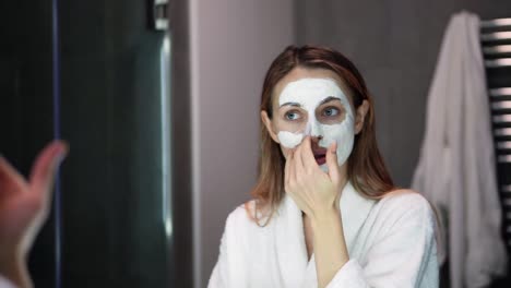 Blonde-woman-in-bathrobe-putting-white-mask-for-moisturizing-using-fingers,-slowmo