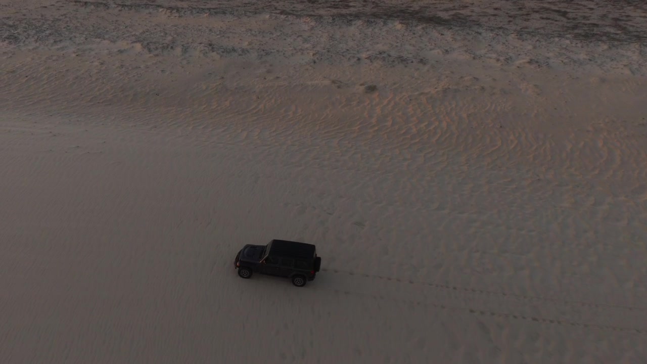 Jeep che attraversa un terreno misto a Guríu, Ceará, Brasile, dove piane di marea scure e compatte incontrano morbide dune di sabbia elevate, creando un forte contrasto nella texture della superficie, ripresa aerea ampia e alta con drone in tracking.