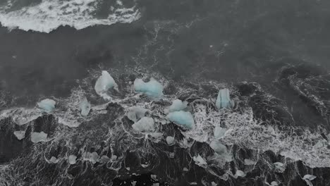 Zeitlupenschwenk-Drohnenaufnahme-Von-Wellen,-Die-Gegen-Eisberge-Am-Schwarzen-Sandstrand-Krachen