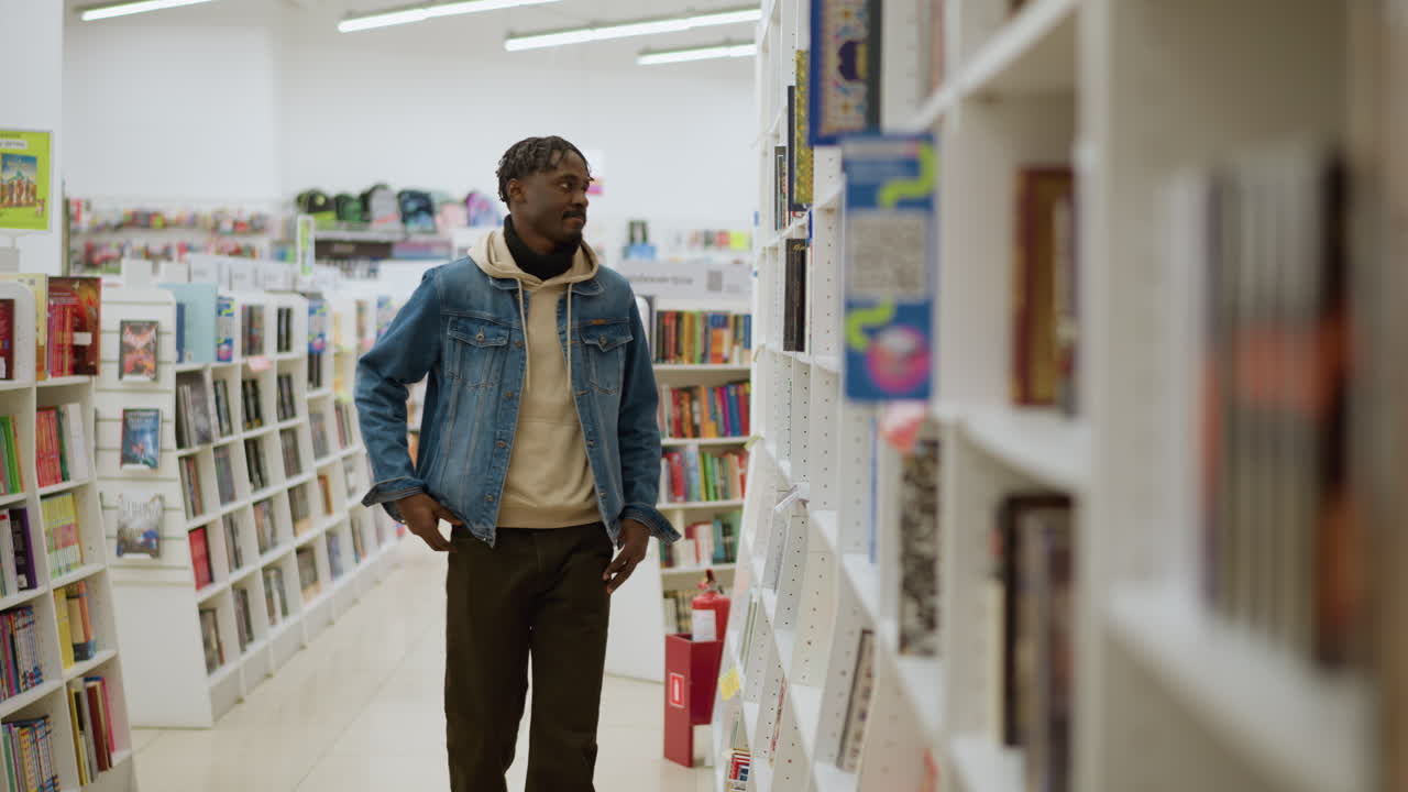 Young man walking between bookshelves in modern library, casually browsing colorful books, focused on selection, bright indoor lighting, wearing denim jacket and hoodie