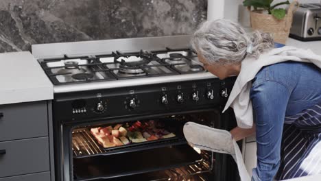 happy senior caucasian woman taking out baked vegetables from oven in kitchen, slow motion