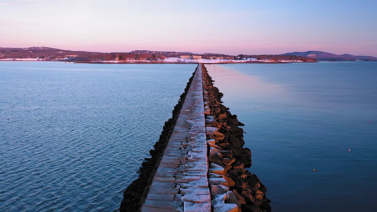 해가 뜰 때 메인 주 록랜드에 있는 눈가루 뿌린 바위 방파제 위로 해안을 향해 날아가는 항공 영상