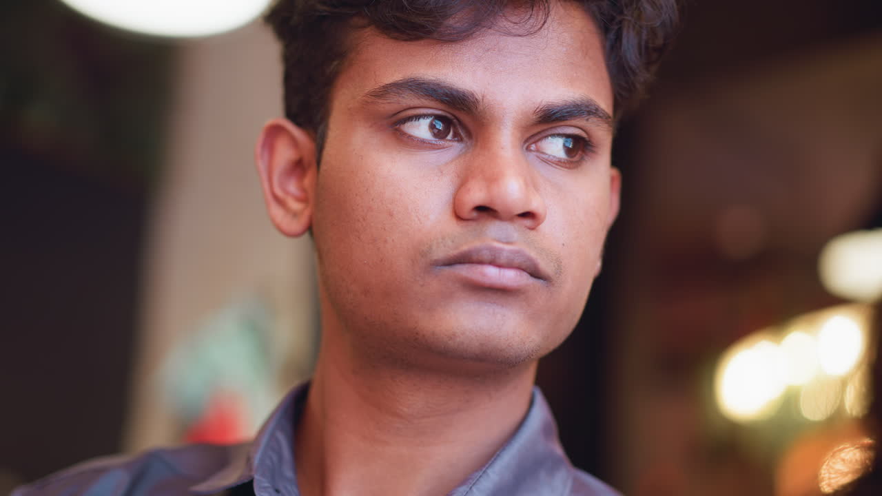 Close-up face of focused young man looking downward in quiet indoor setting, wearing gray shirt, expression showing deep thought or concentration, warm bokeh lights in soft background