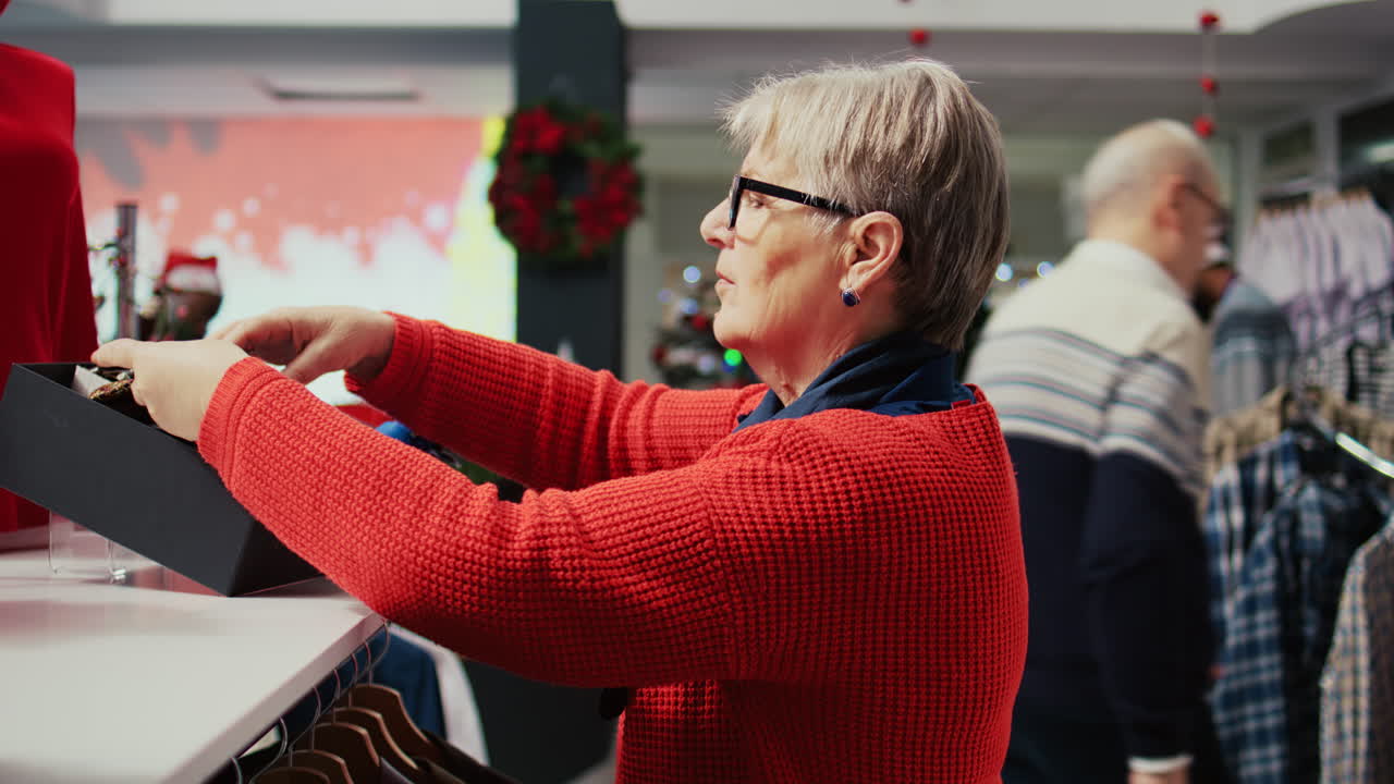 Elderly woman shopping for stylish neckties to gift husband for Christmas during winter holiday season. Senior customer in xmas decorated clothing store looking for presents