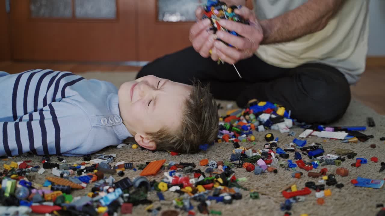 Little boy lying on the floor and the man pours pieces of plastic lego bricks on the kid's face. Father and son in the living-room playing with lego blocks. Family and childhood concept.