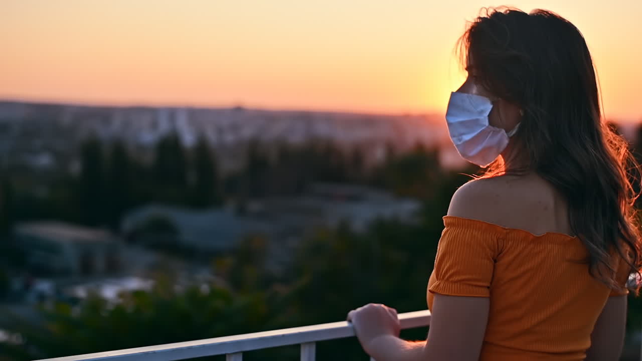 Young woman in carrot blouse looking at the city and enjoying orange sunset from the observation deck in the evening. Wearing protective medical mask. Rear view. Corona Virus idea