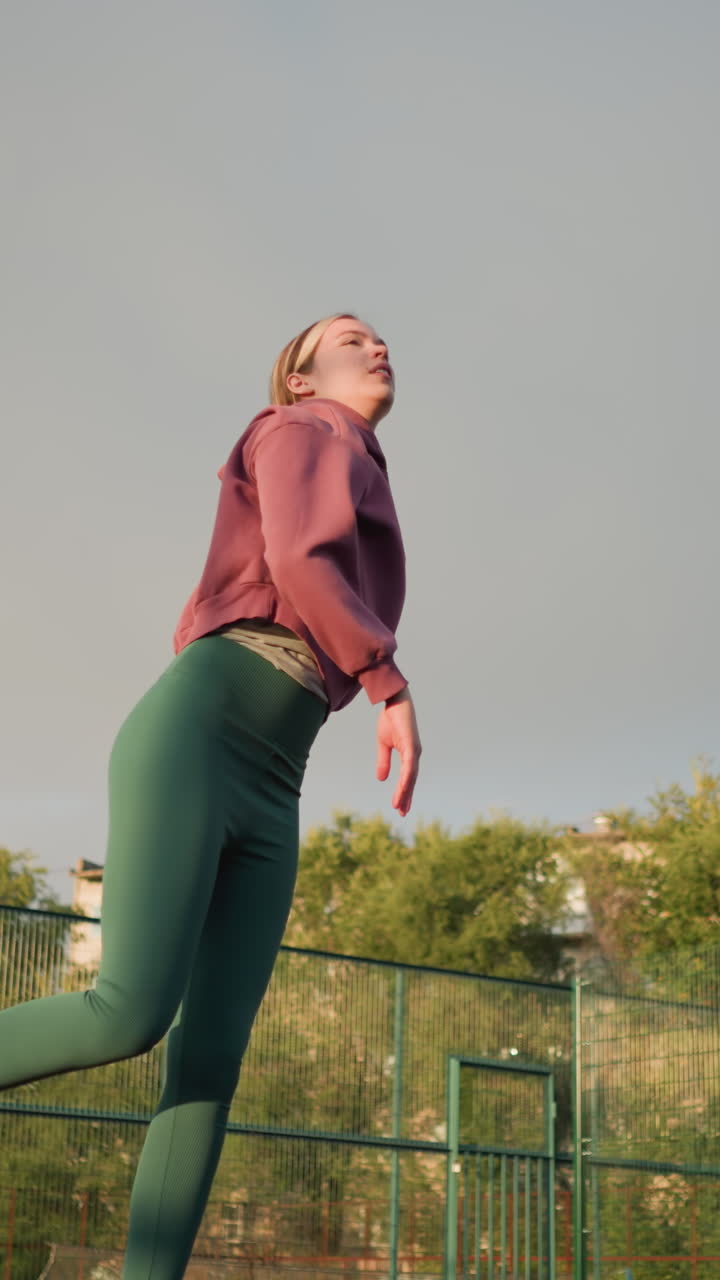 dama en la cancha al aire libre salta, lanza la pelota de voleibol hacia arriba, espera que aterrice, luego la atrapa, aro de baloncesto visible en el fondo con personas sentadas, vegetación y edificios que rodean el área