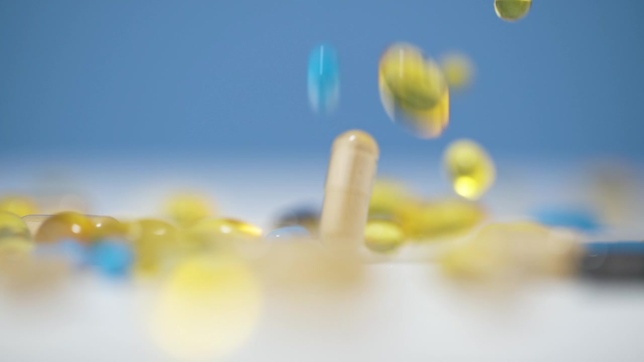 Pillole in capsule mediche gialle e blu che cadono su una superficie bianca con sfondo sfocato.