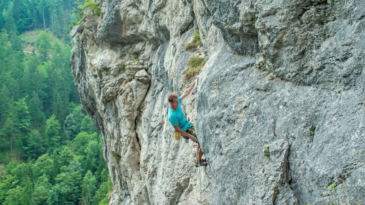 Caucasian man climbing up mountain, takes rope and clips in to side of mountain