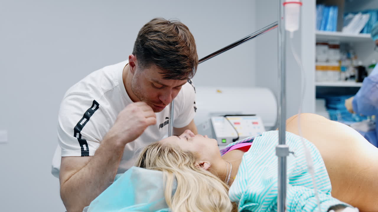 Man bent over his pregnant wife lying on the bed. Woman is prepared for the c-section operation and husband soothes her.