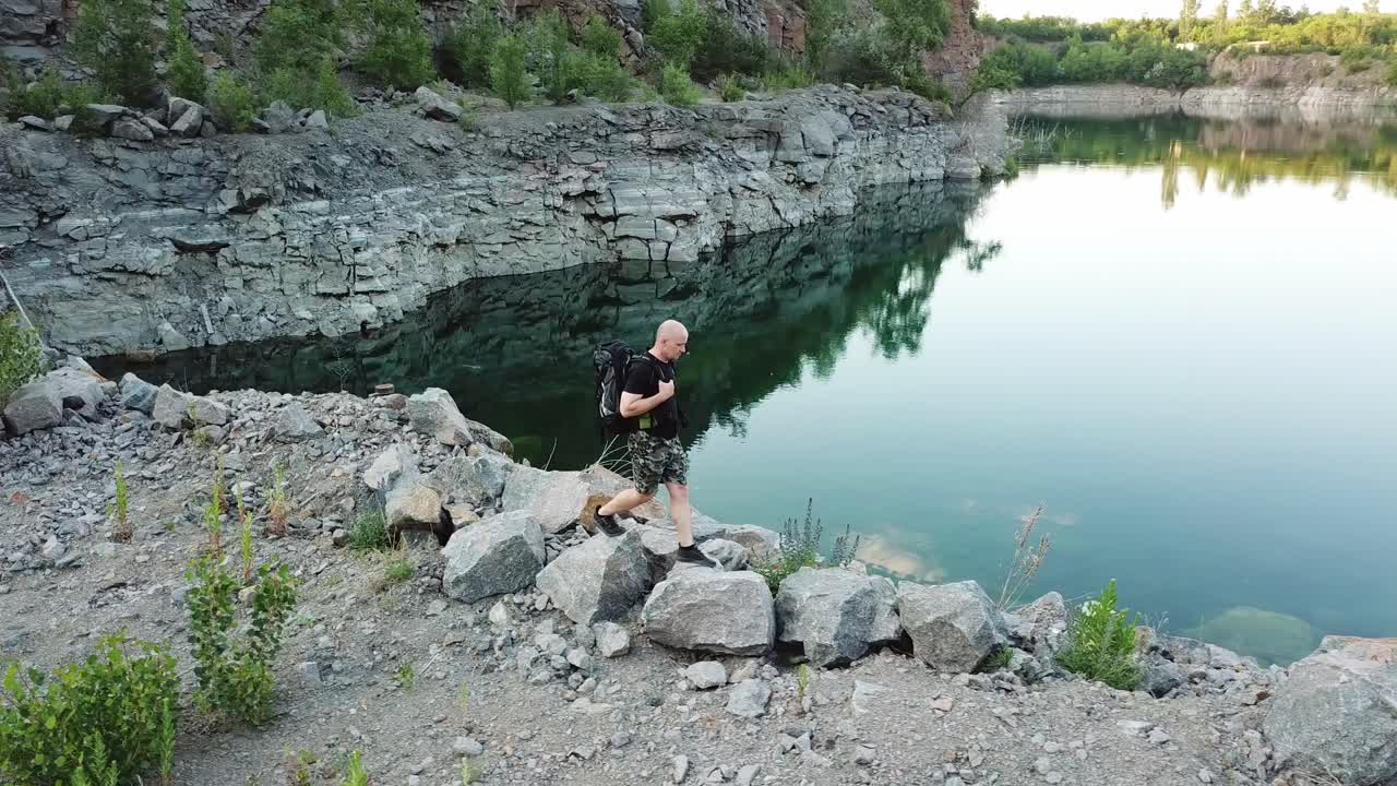 A traveler in a black T-shirt with a backpack on his shoulders walks along the waterside of the lake near the quarry with stones and trees. Wonderful summer landscape.