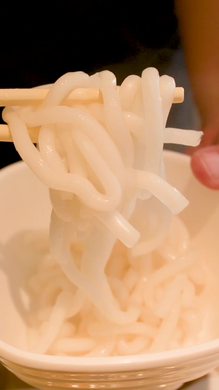 Primo piano di mani che usano bacchette di legno per sollevare spessi noodles bianchi da una ciotola.