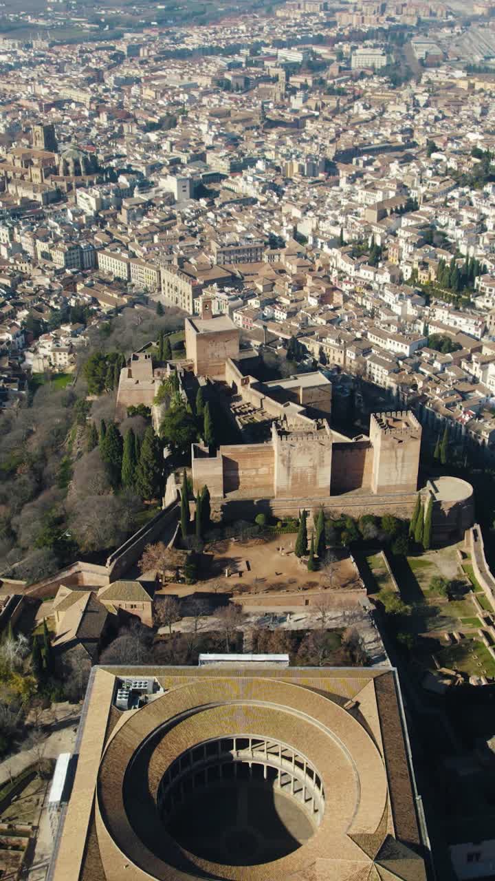 Drone verticale in volo all'indietro sopra il Palazzo di Carlo V all'interno della cittadella dell'Alhambra, rivelando lo skyline di Granada sullo sfondo.