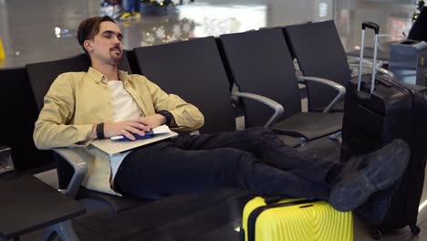 Un-Hombre-Barbudo-Con-Camisa-Amarilla-Durmiendo-Sosteniendo-Las-Piernas-En-La-Maleta-Amarilla,-Sosteniendo-Sus-Cosas-En-El-Salón-Del-Aeropuerto-Mientras-Estaba-Sentado