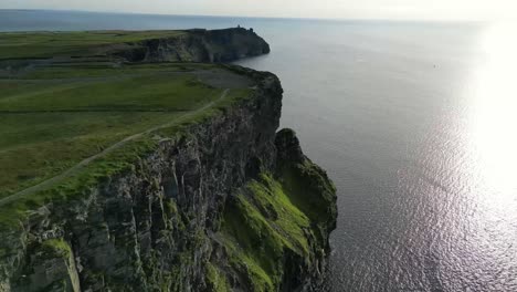 Drone-shot-panning-left-to-right-along-the-Cliffs-of-Moher-at-sunset,-while-hikers-move-along-the-cliffs