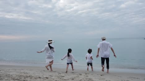 rear view happy asian family on summer vacation father, mother, son and daughter running into sea together on the beach in the morning time, sunrise. holiday and travel concept. slow motion