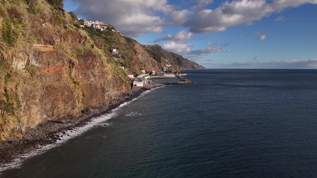 Video drone mozzafiato scorre lungo le aspre scogliere marine di Madeira verso un tranquillo villaggio costiero adagiato sotto colline vulcaniche, con un'illuminazione drammatica e ombreggiata e nuvole sparse sopra