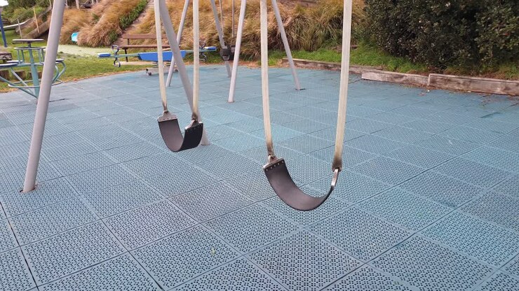 Kid's empty swings swaying and swinging in the breeze in empty playground with safety matting, close up of swings