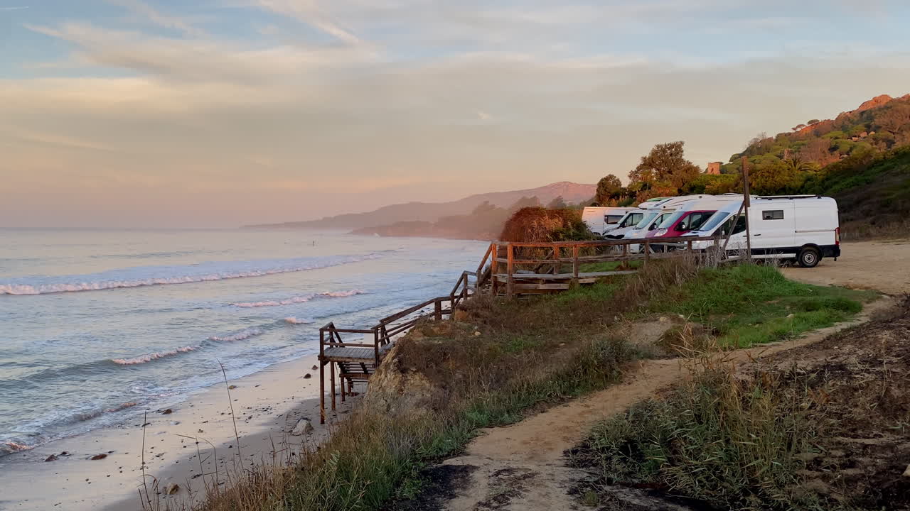 A raised wooden platform stands above the sand at Hurricane, Tarifa, facing soft evening waves and a hazy pastel horizon with camper vans in the shhot