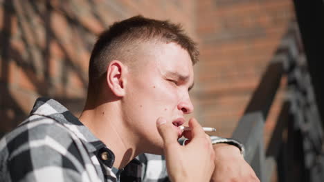 young boy with spots on face smoking cigarette while leaning on iron railing under bright sunlight casting shadows on brick wall in background with soft blur and thoughtful facial expression