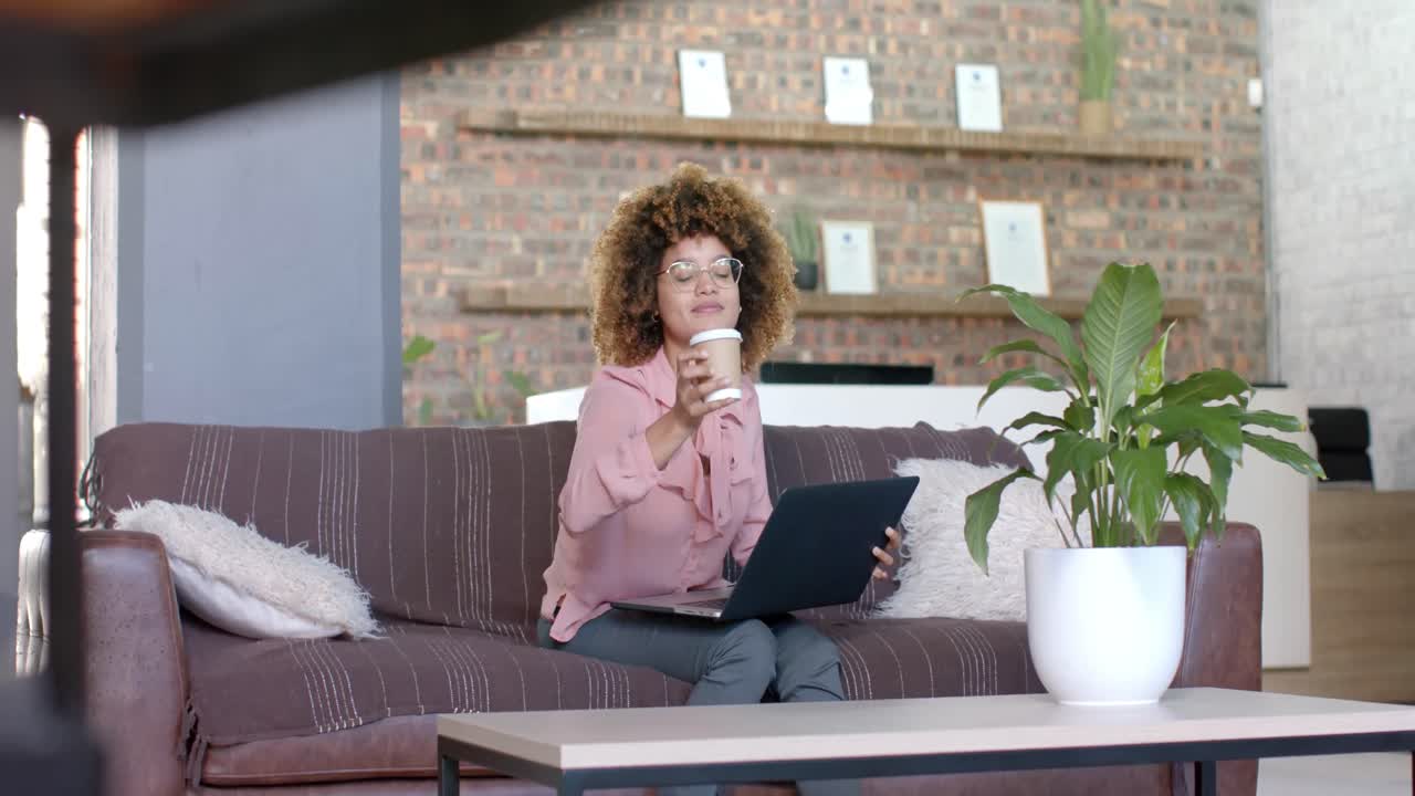 Happy biracial casual businesswoman drinking coffee and using laptop on couch in office, slow motion