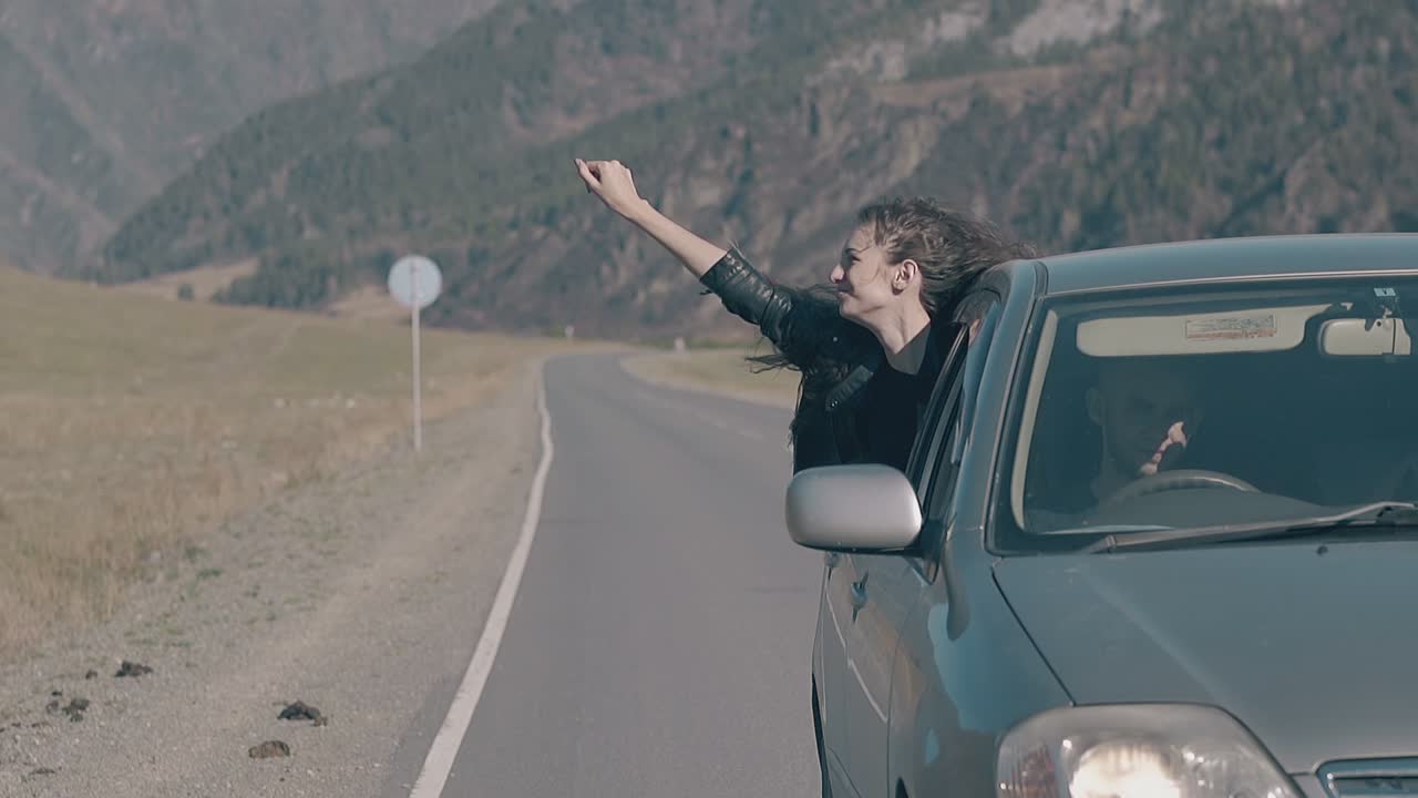 молодая девушка махает рукой и наслаждается жизнью, откидываясь от машины.