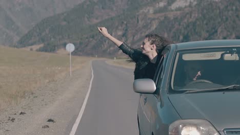 young girl waves hand and enjoys life leaning out of car