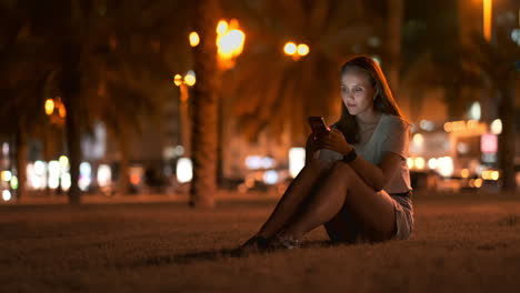 At-night-a-young-girl-sits-on-the-grass-in-a-big-city-and-looks-into-the-smartphone-screen-writes-a-message-and-maintains-her-personal-blog.