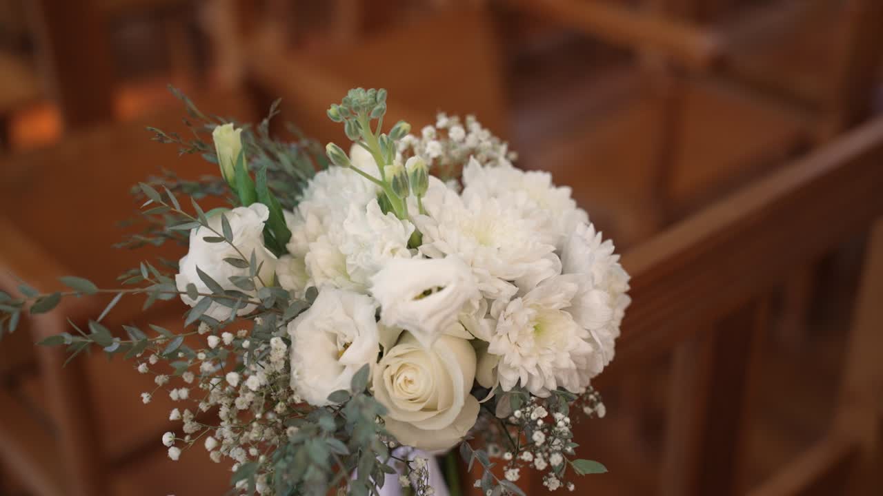 A close-up view of a bouquet full of white flowers is shown. The event takes place inside a rustic hall with wooden chairs arranged for guests