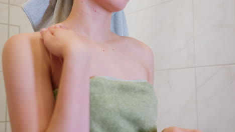 close-up view of woman wrapped in light green towel applying cream on her neck, she is in the bathroom, focusing on moisturizing and enhancing skin care with a gentle, soothing motion