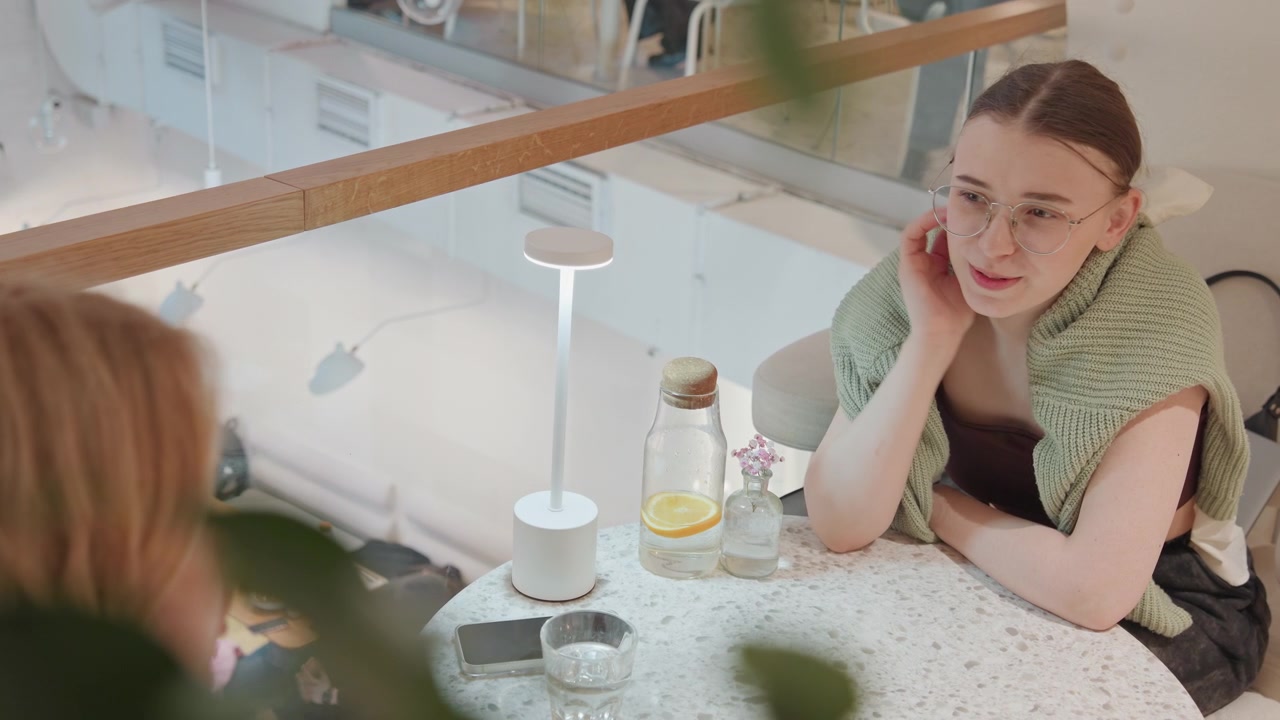 Primo piano di una giovane donna con gli occhiali che riposa a un tavolo di un caffè moderno, impegnata in una conversazione. La donna indossa un maglione verde lavorato a maglia sopra abiti scuri; sul piano in marmo ci sono due bottiglie di vetro, una