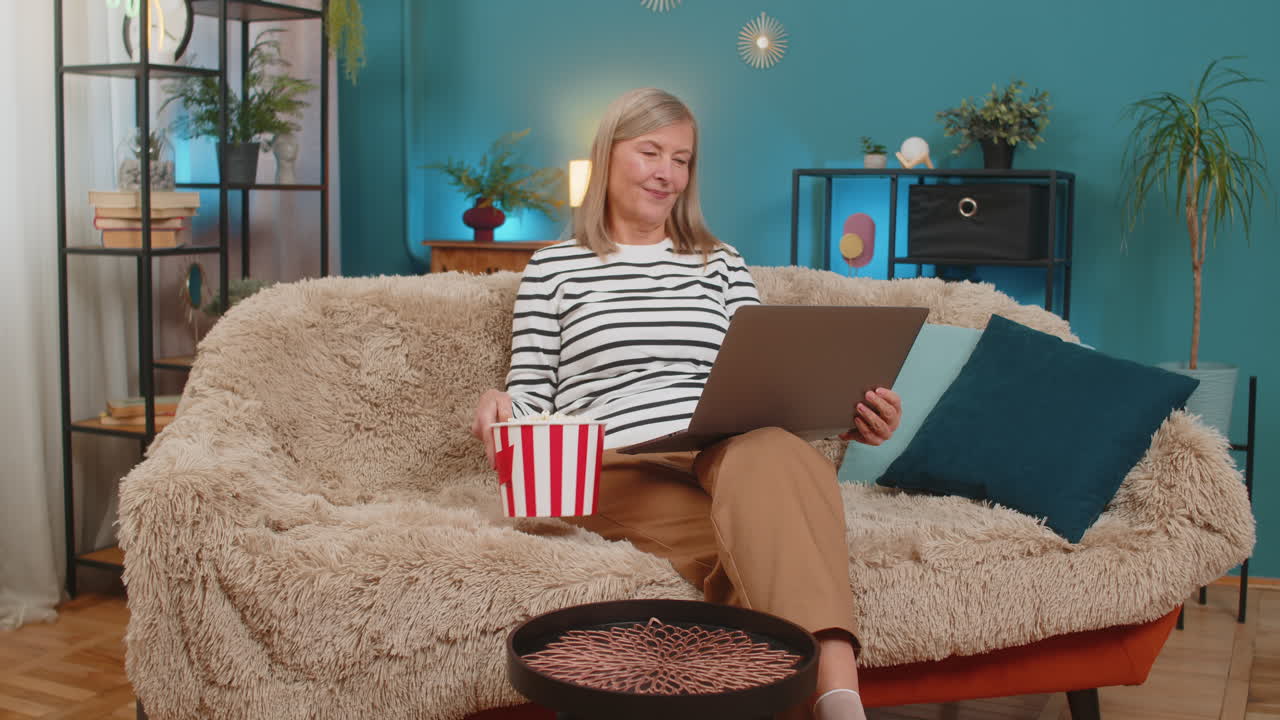 Mature woman placing popcorn bucket on table setting up laptop to watch series relaxed at home