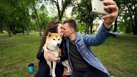 happy couple is taking selfie in the park kissing and looking at smartphone, while young woman is holding and caressing cute puppy shiba inu breed.