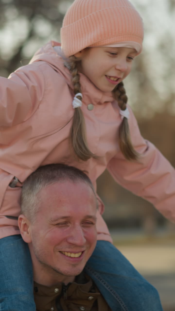 Cheerful father and daughter moment with the girl in a pink cap and jacket, arms spread wide, sitting on his shoulders