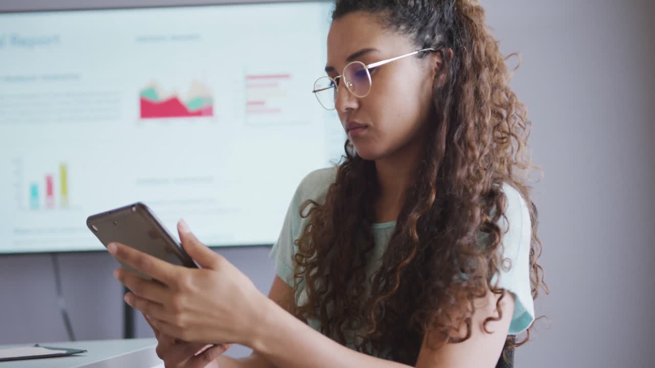 Biracial businesswoman wearing glasses using tablet in meeting room with screen in background