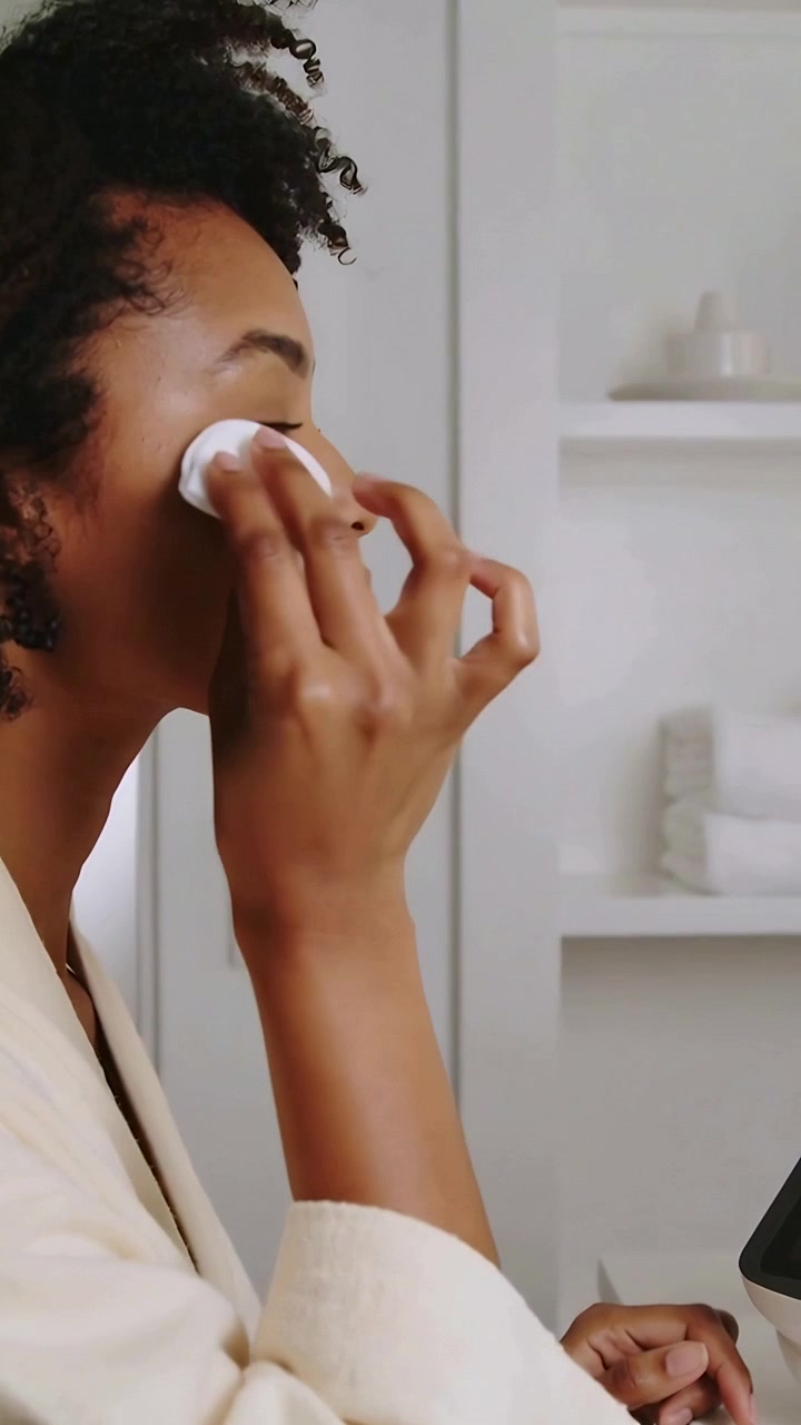 Vertical video: Holding cotton pad, woman in robe wiping face at vanity, removing makeup