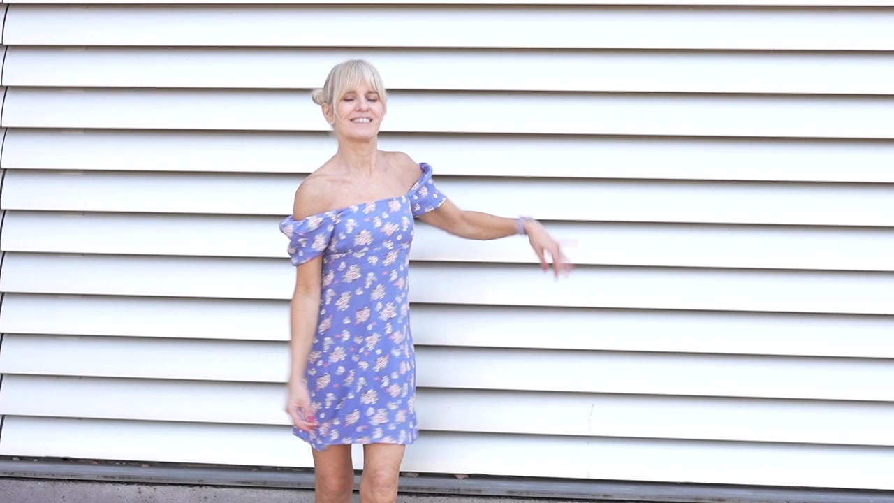 Una donna in abito floreale posa all'aperto davanti a un muro bianco, emanando eleganza e sicurezza.