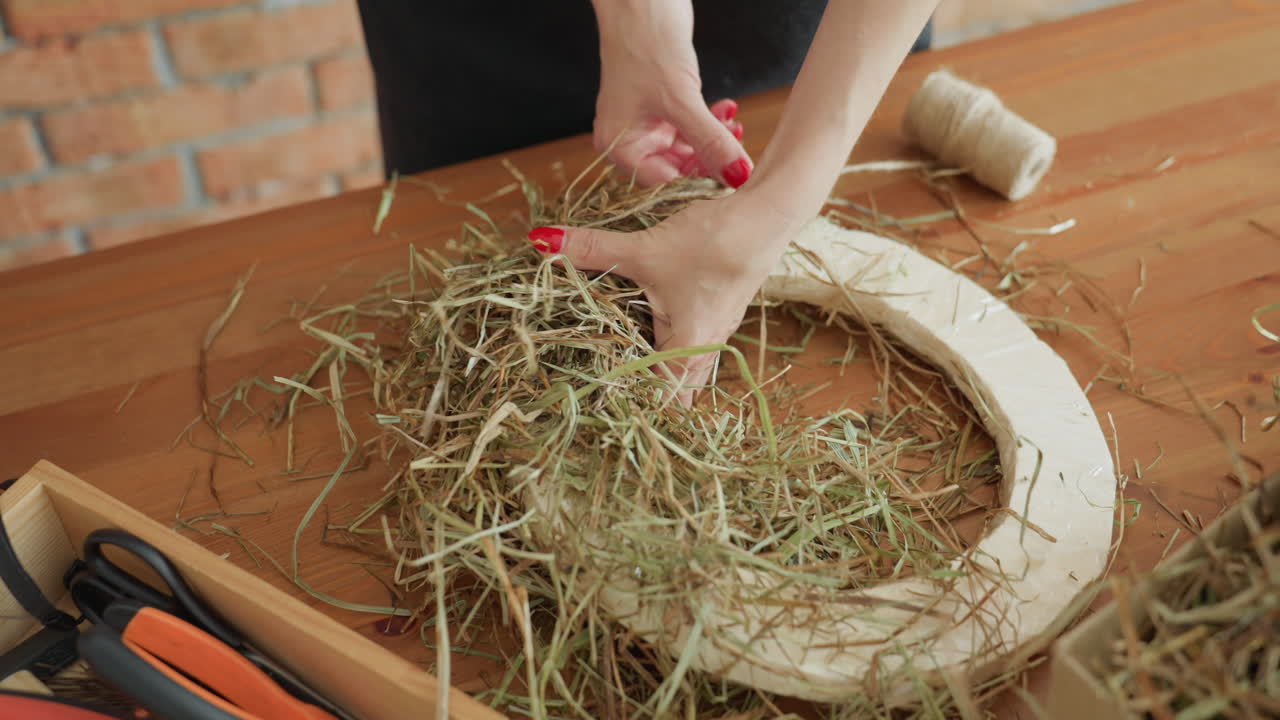 Decorator shaping dried hay on circular base using hands with red nails, crafting rustic wreath with twine spool and tools on wooden table in creative workspace focused on handmade decoration process